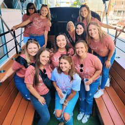Smiling bachelorette group on an open-air party trolley in downtown, bride in a light blue 'Bride' tee with friends in matching pink shirts and jeans holding tumblers.