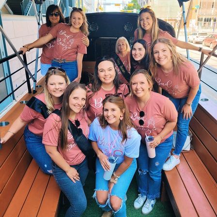 Smiling bachelorette group on an open-air party trolley in downtown, bride in a light blue 'Bride' tee with friends in matching pink shirts and jeans holding tumblers.