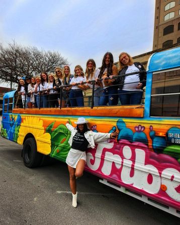 Group of women smiling on a colorful graffiti-style party bus parked on a downtown street, one woman in a white hat and boots posing in front.