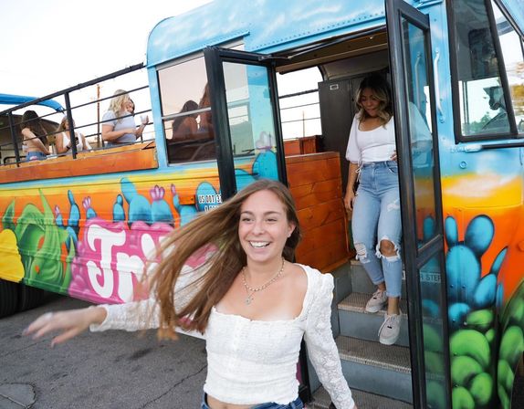 Smiling young woman rushing off a colorful graffiti-painted party bus on a sunny street, friend following down the steps and passengers seated inside