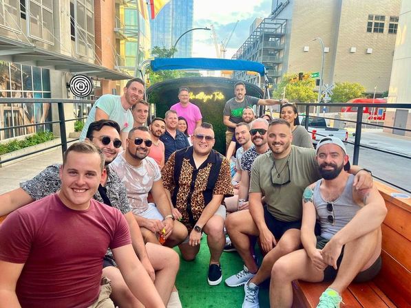 Smiling group of about sixteen adults seated on wooden benches of an open-air party wagon enjoying a sunny day on a downtown city street with modern buildings and traffic in the background.