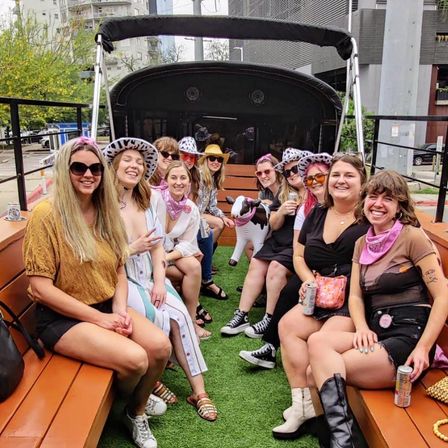 Group of friends smiling on a covered outdoor party wagon in a city, seated on wooden benches over artificial turf, wearing cowboy hats and pink bandanas, holding drinks and an inflatable cow.
