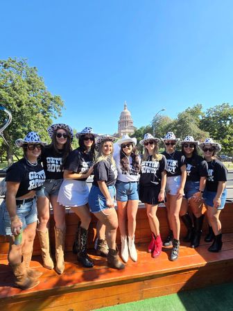 Group of women in matching black "Gettin Rowdy" shirts, spotted cowgirl hats and cowboy boots posing on a wooden platform with the Texas State Capitol dome and clear blue sky in Austin, TX.