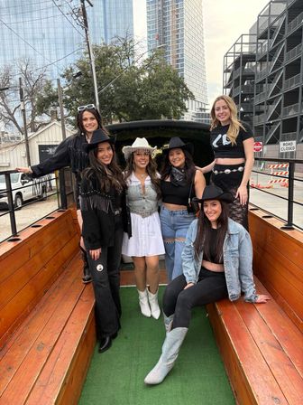 Smiling group of six friends in cowgirl hats, boots, and fringe outfits posing on a wooden outdoor wagon with a downtown city skyline and parking structure in the background.