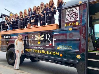Group of women smiling and posing on an open-top party bus parked on a downtown city street, one woman in white standing on the sidewalk in front — festive daytime urban tour scene.