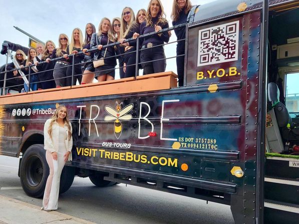 Group of women smiling and posing on an open-top party bus parked on a downtown city street, one woman in white standing on the sidewalk in front — festive daytime urban tour scene.