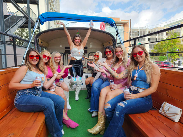 Group of eight women in colorful festival outfits and heart-shaped sunglasses partying on a wooden-seated pedal pub trolley in a downtown urban setting, one standing with arms raised wearing shiny holographic boots.