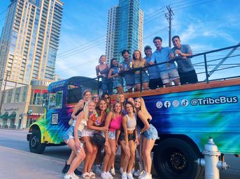 Group of smiling young friends in casual summer outfits posing with drinks by a colorful party bus on a downtown street lined with high‑rise buildings at sunset.