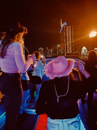 Neon-lit rooftop boat party at night with friends dancing, a pink fuzzy cowboy hat in the foreground and a glowing downtown skyline of high-rise buildings in the background.