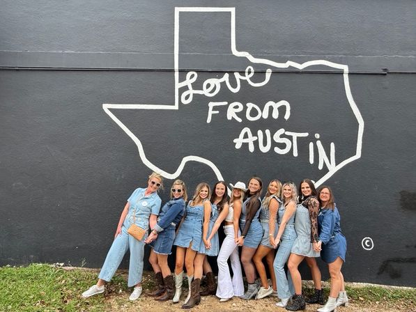 Smiling group of women in denim outfits and cowboy boots posing in front of a black mural of the Texas outline that reads "Love From Austin" — popular Austin, Texas photo spot.