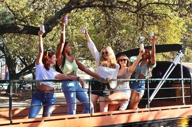 Five friends on a sunlit riverside boat deck raising canned drinks and laughing under shady trees, wearing casual summer outfits.