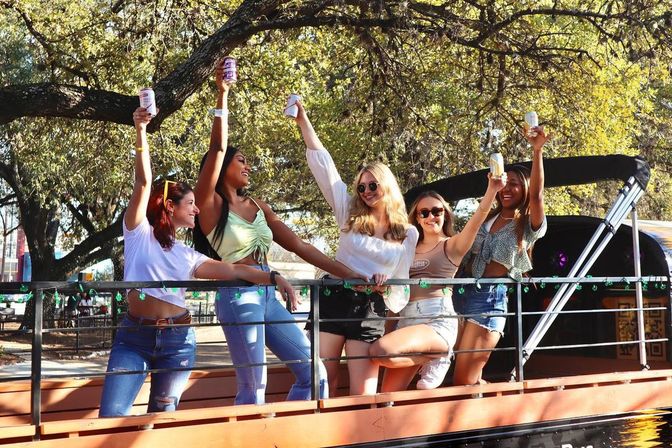 Five friends on a sunlit riverside boat deck raising canned drinks and laughing under shady trees, wearing casual summer outfits.