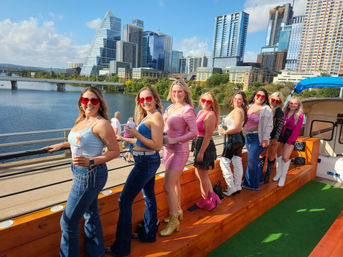 Group of women in colorful outfits and heart-shaped sunglasses posing on a boat deck with drinks, Austin, Texas skyline and river in the background on a sunny day.