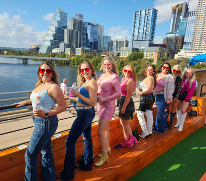 Group of women in colorful outfits and heart-shaped sunglasses posing on a boat deck with drinks, Austin, Texas skyline and river in the background on a sunny day.