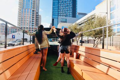 Two friends celebrating on an open-top party bus in a downtown urban setting, raising drinks toward a backdrop of glass high-rise buildings.