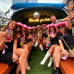 Group of friends on an outdoor party boat wearing pink cowboy hats and bandanas, holding colorful drinks under a disco ball and neon “Vibe with your tribe” sign — lively bachelorette-style celebration in a city setting.