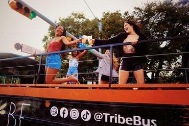 Four friends laughing and clinking drinks on an open-top party bus cruising a downtown city street with trees and traffic lights