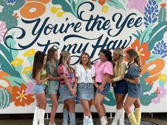 Seven women in denim skirts and cowboy boots posing and laughing in front of a colorful floral mural that reads You're the Yee to my Haw.