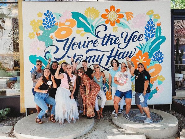 Lively group photo of friends posing and cheering in front of a colorful floral-and-cactus wall mural that reads "You're the Yee to my Haw" — outdoor street-art backdrop with a playful, celebratory vibe.