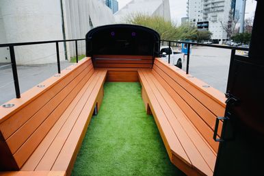 Open-air trailer interior with long orange wooden benches on either side, green artificial-turf aisle, metal cup holders and railing, parked on a downtown urban street with apartment buildings visible.