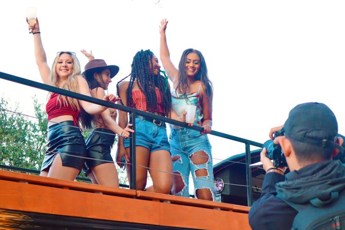 Four friends posing and dancing on a sunny rooftop balcony at an outdoor party, one holding a drink while a photographer snaps photos.