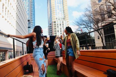Four friends laughing and sipping drinks on a downtown rooftop patio with wooden benches and high-rise buildings under a bright blue sky.