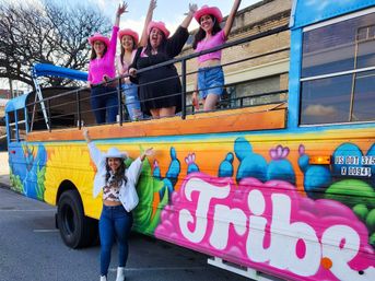 Five people wearing pink cowboy hats cheer on and around a colorful painted party bus with bright cactus mural and bold graffiti-style lettering, parked on a sunny street.