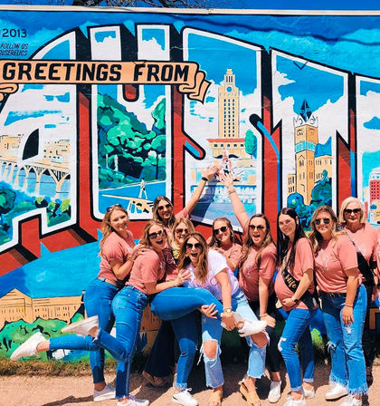 Group of friends in matching pink shirts and jeans laughing and posing playfully in front of a bright “Greetings From” postcard-style city mural outdoors.