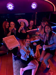 Group of friends laughing and eating pizza inside a neon-lit party bus at night, some wearing playful hats and holding pizza boxes.
