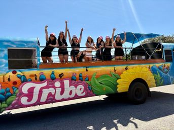 Group of eight women in black outfits and pink caps waving from an open-top colorful party bus with a floral and music mural on a sunny day.