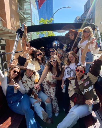 Group of friends partying on an open-top downtown party vehicle, smiling and raising canned drinks, wearing sunglasses and trendy outfits under a hanging disco ball on a sunny city street.