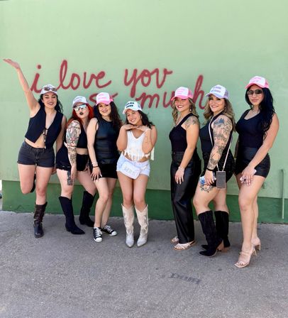 Seven friends wearing matching pink trucker hats pose playfully in front of a pale green outdoor mural painted with red “i love you so much” script; most wear black outfits while one wears a white fringe top and silver cowboy boots, showing tattoos, smiles, and varied boots and heels.