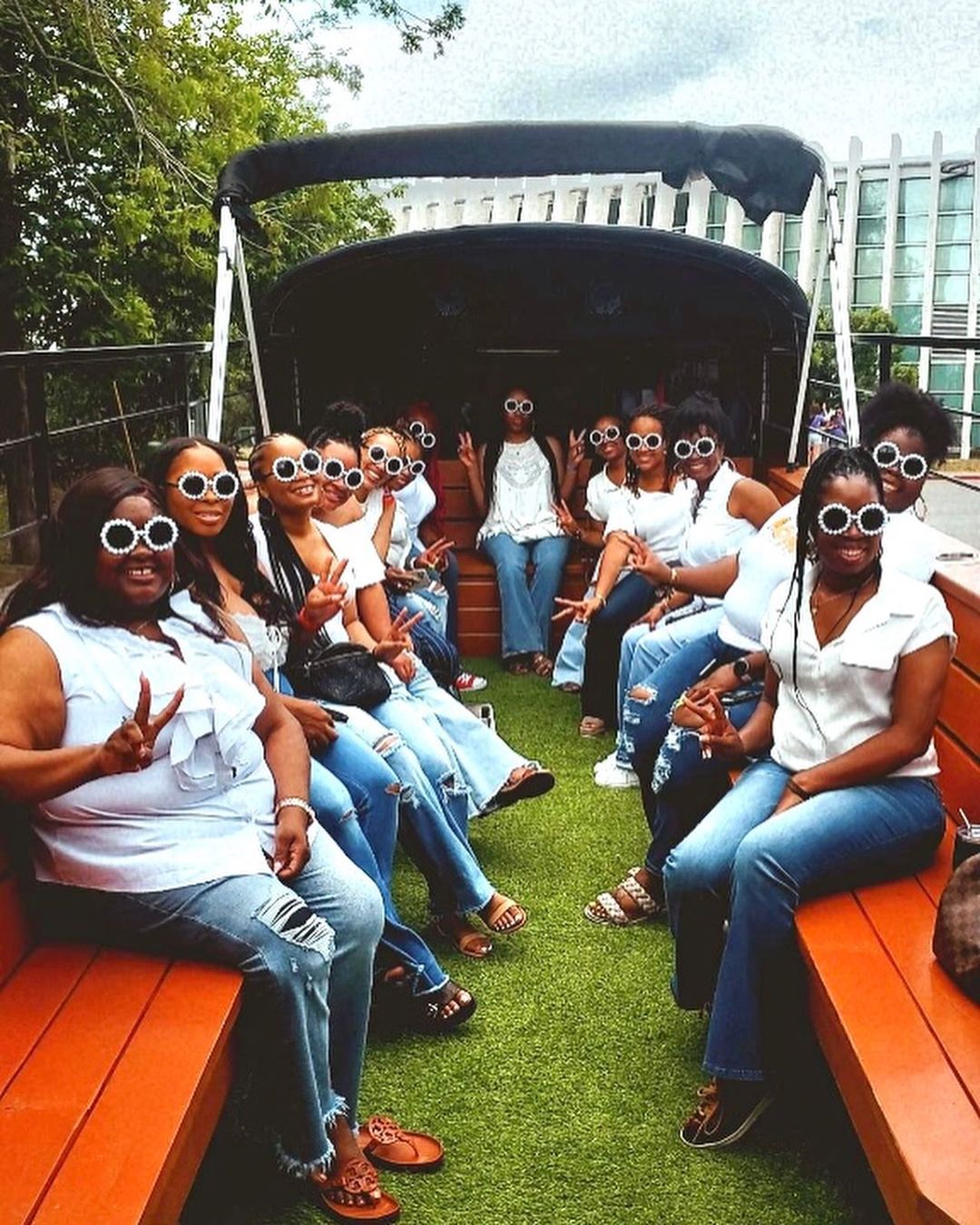 Festive group of women in white tops and jeans wearing matching round white sunglasses, seated on wooden benches inside an open-air party bus with green turf floor, smiling and flashing peace signs against a tree and glass-building backdrop.