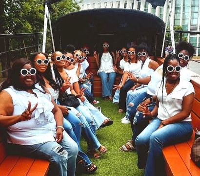 Festive group of women in white tops and jeans wearing matching round white sunglasses, seated on wooden benches inside an open-air party bus with green turf floor, smiling and flashing peace signs against a tree and glass-building backdrop.