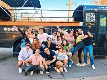 Group of friends posing cheerfully in front of a black party bus with rooftop deck, holding drinks and flashing peace signs on a sunny street.