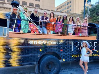 Group of friends in colorful retro outfits and sunglasses posing on an open-top party bus parked on a downtown street, with a bride-to-be in a white fringe dress and veil standing beside the bus.