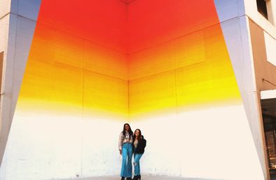 Two smiling friends in jeans pose at the base of a large triangular gradient mural (red to yellow to white) on an urban corner wall.