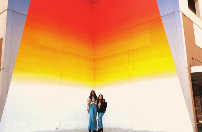 Two smiling friends in jeans pose at the base of a large triangular gradient mural (red to yellow to white) on an urban corner wall.