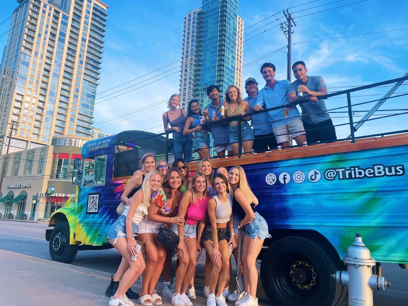 Smiling group of friends posing with drinks on a colorful open-top party bus parked in a downtown street against a backdrop of glass high-rise buildings and blue sky.