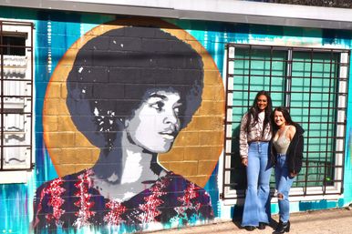 Two friends pose and smile beside a bold street-art mural of a woman with an afro and gold halo painted on a turquoise cinderblock wall with barred windows