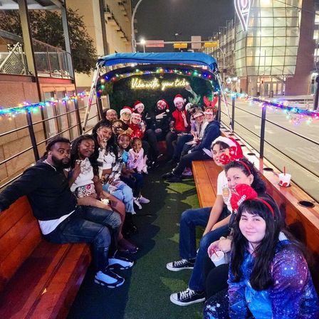 Festive group of adults and kids in Santa hats and reindeer antlers riding a light‑decorated open-air holiday trolley along a downtown street at night.