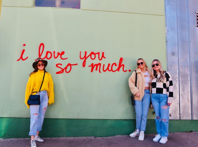 Three friends posing at an urban photo spot in front of a mint-green wall with red cursive graffiti reading “i love you so much,” wearing casual denim and sunglasses.