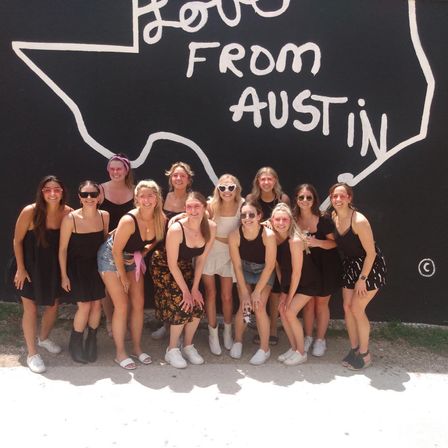 Smiling group of women in summer outfits posing on a sunny sidewalk in front of a bold black-and-white “Love From Austin” Texas mural.