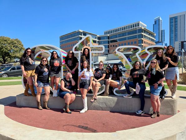 Group of women in matching graphic tees and boots posing on and around a large colorful "ATX" sculpture in downtown Austin, Texas, with modern buildings and a bright blue sky behind them.