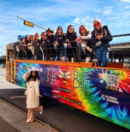 Group of women in sunglasses leaning on the railing of a brightly tie-dye open-top party bus parked on a sunny city street, while a woman in a white coat and sequined skirt poses on the sidewalk.
