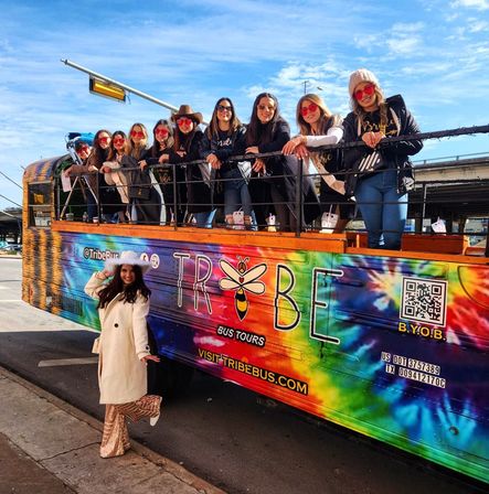 Group of women in sunglasses leaning on the railing of a brightly tie-dye open-top party bus parked on a sunny city street, while a woman in a white coat and sequined skirt poses on the sidewalk.