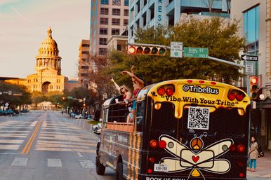 Open-air party bus with passengers waving down a quiet downtown Austin, Texas street toward the sunlit state capitol dome at golden hour, colorful painted rear with QR code, 8th Street sign and traffic lights overhead.