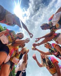 Low-angle shot of friends in colorful tie-dye shirts and fanny packs toasting canned drinks under a bright, sunny summer sky at an outdoor celebration.