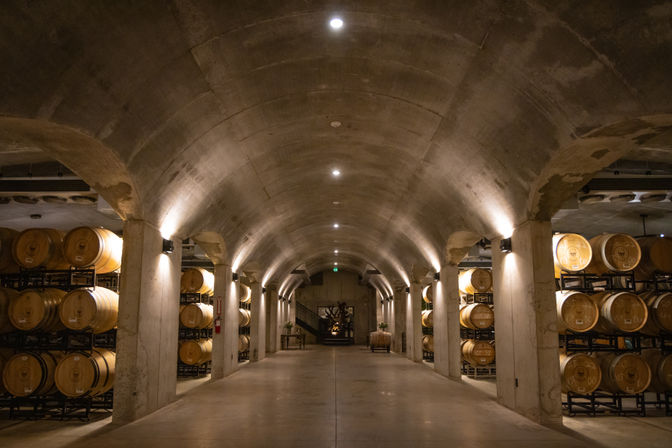 Underground winery cellar lined with stacked oak barrels under an arched concrete ceiling, warm uplighting and a wide central aisle
