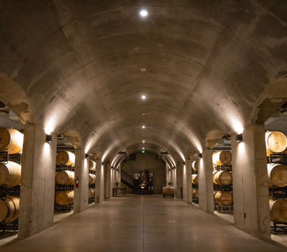 Underground winery cellar lined with stacked oak barrels under an arched concrete ceiling, warm uplighting and a wide central aisle